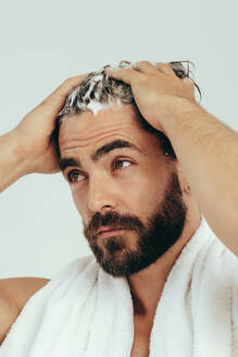 Ein gut aussehender junger Mann trägt Shampoo auf sein Haar auf und bereitet sich auf eine gründliche, erfrischende Wäsche vor. Ein Mann, der sich mit einem Badetuch auf den Schultern auf seine Pflege und Selbstfürsorge vorbereitet. - JLPSF29978