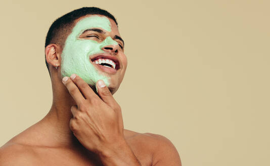 Ein glücklicher junger Mann, der seine Haut mit einer Gesichtsmaske verwöhnt. Der Mann lässt die pflegenden Inhaltsstoffe eines Gesichtsreinigers als Teil seiner Hautpflegeroutine auf sein Gesicht einwirken. - JLPSF29987