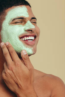 Ein junger Mann lächelt, als er sich eine grüne Gesichtsmaske gönnt. Ein glücklicher junger Mann, der seine Haut mit einem feuchtigkeitsspendenden Gesichtsreiniger pflegt und verwöhnt. - JLPSF29989