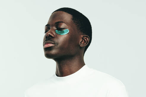 Black man indulging into some under eye care with a hydrogel patch. Standing in a studio with his yes closed, he absorbs the benefits of the beauty mask on his healthy and glowing melanin skin. - JLPSF29991