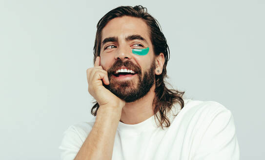 Happy young man wearing an undereye mask in a studio, pampering his face with the gel patch to reduce circles under the eyes. Handsome young man practicing an anti-aging self-care routine. - JLPSF29999