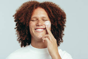 Man with a curly afro taking care of his skin using a moisturizing facial cream. Handsome young man practicing his skincare routine with a big smile, enjoying the grooming part of lifestyle. - JLPSF30005