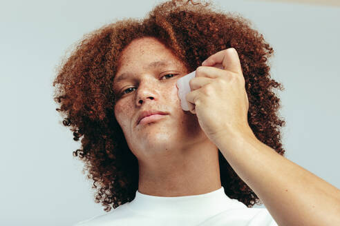 Junger Mann, der sein Gesicht mit einem Gua Sha-Stein massiert, um Schwellungen zu reduzieren und sein Aussehen zu verbessern. Mann mit Sommersprossen, der seine einzigartige Haut mit einer gesunden Hautpflege verwöhnt. - JLPSF30007