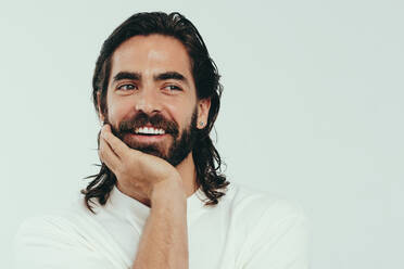 Handsome young man smiling in a studio, proudly wearing his beautiful beard and long hair, as well as his glowing and youthful skin. Happy caucasian male embracing grooming and self-care. - JLPSF30034