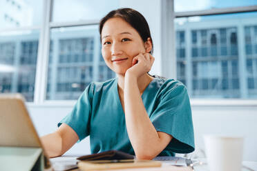 Krankenpflegeschülerin schaut in die Kamera, während sie in einer Bibliothek sitzt, umgeben von medizinischen Lehrbüchern und Studienmaterial. Junge asiatische Frau, die sich auf eine Prüfung in der Krankenpflegeschule vorbereitet. - JLPPF01666