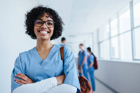 Young female nursing student stands in a medical clinic dressed in scrubs, smiling confidently at the camera. Happy woman in her 20's showing excitement for her medical internship in a hospital. - JLPPF01678