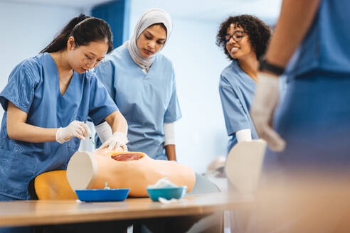 Asian nursing student administers an injection on a patient simulator during training in nursing school. Group of female students in scrubs practicing nursing during a healthcare simulation. - JLPPF01691