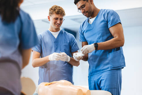 Male students practicing nursing during simulation-based training. Two young men, dressed in scrubs, stand together and prepare to inject a patient simulator in a teaching hospital. - JLPPF01694
