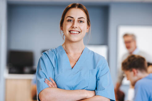 Happy medical student standing in her scrubs with crossed arms, studying studying healthcare education at university. Caucasian woman in her 20s pursuing a career in medicine. - JLPPF01723