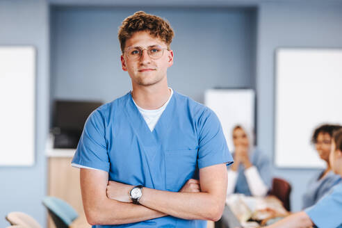 Caucasian medical student stands in a training ward, wearing scrubs and looking at the camera. Young man studying to be a healthcare professional through extensive medical training. - JLPPF01724