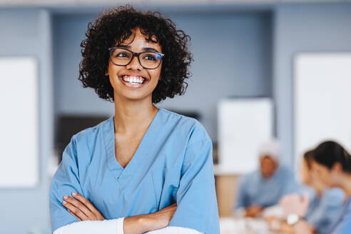 Female medical student in her 20s stands and smiles in scrubs, proudly studying medicine at university. Young woman with curly hair doing her medical training at a teaching hospital. - JLPPF01725