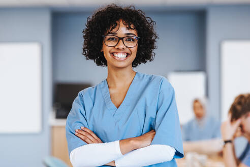 Young female medical student stands in a hospital training ward, smiling at the camera with her arms crossed. Woman in her 20s studying to become a healthcare professional. - JLPPF01726
