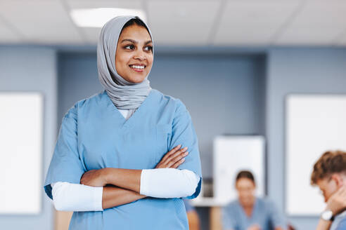 Young female healthcare student wearing a hijab smiles while standing in a medical school, studying for her upcoming residency. Muslim woman in her 20s pursuing a medical qualification. - JLPPF01727