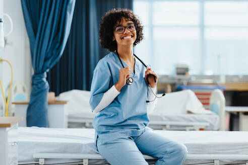 Happy young woman in scrubs smiles at the camera as she sits on a bed during her medical residency at a teaching hospital, furthering her education and healthcare training. - JLPPF01729