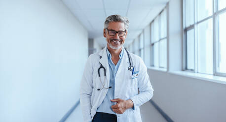 Portrait of a senior medical professional at work. Mature and happy doctor with grey hair stands in a hospital corridor wearing a lab coat and a stethoscope, ready to provide expert healthcare. - JLPPF01742