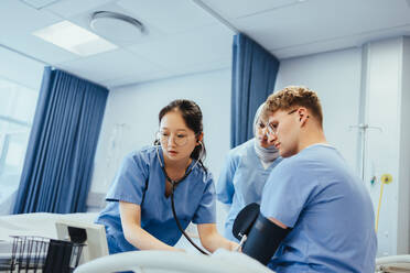 Eine asiatische Medizinstudentin beobachtet ein Blutdruckmessgerät, während sie in einem Krankenhaus die Blutwerte ihrer Klassenkameradin misst. Multiethnische Medizinstudenten üben die Messung der Vitalparameter eines Patienten in der medizinischen Fakultät. - JLPPF01771