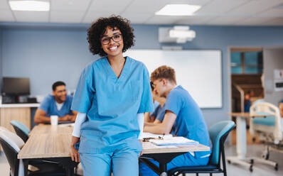 Happy nursing student in scrubs looking at the camera as she stands in a teaching hospital, with her medical study group in the background. Young female student going through nursing training. - JLPPF01791