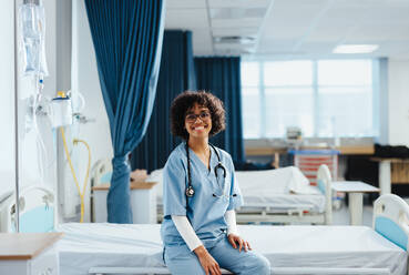 Porträt einer Medizinstudentin, die in einem Universitätskrankenhaus auf einem Bett sitzt, einen medizinischen Kittel trägt und ein Stethoskop um den Hals hat. Die Medizinstudentin lächelt selbstbewusst in die Kamera. - JLPPF01798