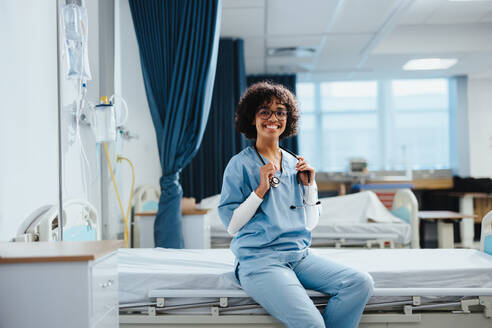 Aspiring medical doctor, a young woman wearing scrubs, smiles at the camera as she sits on a bed in a hospital. Confident female student doing her clinical rotation in medical school. - JLPPF01799