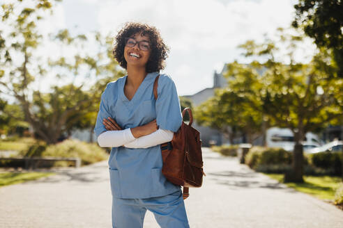 Young, cheerful woman stands outside a university hospital in her doctors scrubs, smiling and happy as she begins the journey of her medical residency. - JLPPF01809