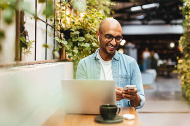 Ein Mann sitzt in einem Café, leger gekleidet und lächelnd, während er mit seinem Smartphone Musik aus einer Streaming-App abspielt. Ein glücklicher junger Mann entspannt sich mit einem Laptop und einer Tasse Kaffee auf dem Tisch in der Nähe. - JLPSF30047
