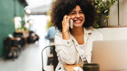 Junge Frau mit lockigem Afro-Haar sitzt in einem Café und lächelt, während sie einen wichtigen Anruf für ihre freiberufliche Tätigkeit tätigt. Glückliche Freiberuflerin, die ein Mobiltelefon benutzt, während sie aus der Ferne arbeitet. - JLPSF30054