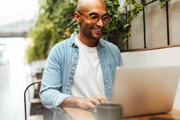 Young black man smiling as he freelances with his laptop at a coworking space, enjoying the ability to work remotely using internet connection. Male digital nomad sitting in a coffee shop. - JLPSF30057