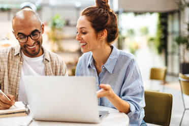 Remote team working together on a project in a coworking space, exchanging ideas and brainstorming. Two happy business people, man and a woman, sitting at a table with a work laptop. - JLPSF30062