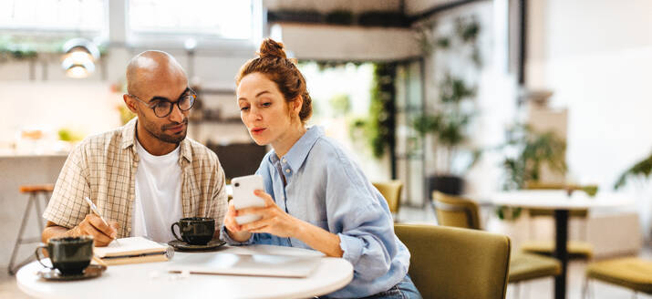 Geschäftsfrau, die ihr Mobiltelefon mit ihrem Kunden teilt, während sie in einer Besprechung die Arbeit bespricht. Kaukasische Unternehmerin, die mit ihrem männlichen Mitarbeiter in einem Café zusammenarbeitet. - JLPSF30064