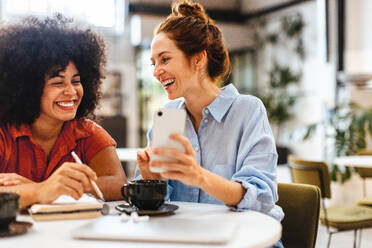 Two female colleagues enjoying a coffee break in their coworking space, reading a client’s text message on a smartphone. Diverse business women smiling as they work together. - JLPSF30066