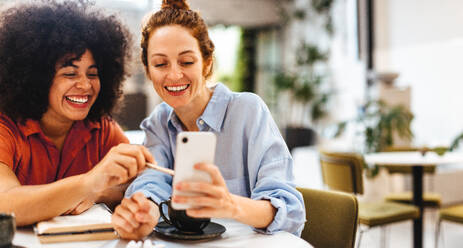 Zwei junge Geschäftsfrauen nutzen ein Smartphone, um während einer Kaffeepause ihre Arbeit zu besprechen und Ideen auszutauschen. Glückliche Unternehmerinnen genießen die Zusammenarbeit in einem Café. - JLPSF30068