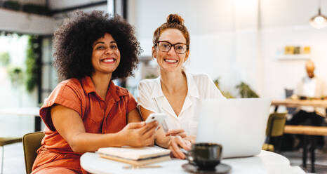 Two female colleagues smiling at the camera, enjoying working together over coffee. Two young women meeting in a a cafe to discuss their collaboration on an important business project. - JLPSF30072