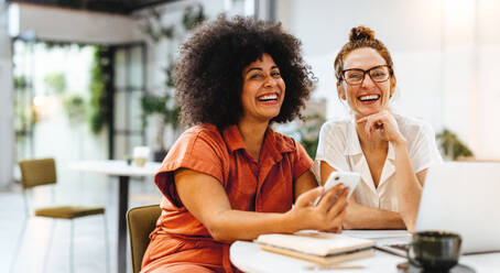 Two successful businesswomen working together on a project in a coffee shop, utilizing their strengths and expertise in a collaboration. Women smiling at the camera during their business lunch. - JLPSF30073