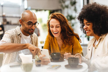 Eine Gruppe von Freunden sitzt in einem Café und nutzt ein Mobiltelefon, um in Verbindung zu bleiben und die neuesten Nachrichten und Updates auszutauschen. Fröhliche junge Leute, die sich bei einem Kaffee unterhalten. - JLPSF30081