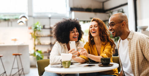 Eine Gruppe fröhlicher Freunde surft in einem belebten Café auf einem Smartphone durch die sozialen Medien und tauscht Fotos und Neuigkeiten aus. Unbeschwerte Freunde haben Spaß, während sie zusammen bei einem Kaffee abhängen. - JLPSF30083