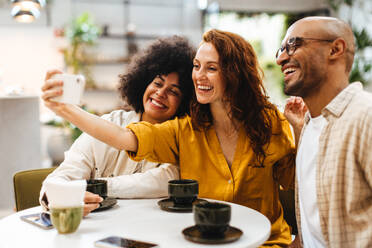 Three friends snap a selfie together in a cozy cafe, capturing a happy moment and creating a cherished memory. Diverse young people having fun together on a relaxed coffee date. - JLPSF30084