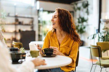 Woman catching up with friends over coffee in a restaurant, chatting and enjoying the lively atmosphere. Happy young, Caucasian woman socializing with a group of people in a cafe. - JLPSF30085