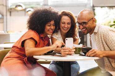 Drei glückliche Freunde machen eine Pause und scrollen durch ihre Lieblings-Social-Media-Apps auf dem Smartphone, lachen und plaudern zwischen zwei Schlucken ihrer heißen Getränke in einem gemütlichen Café. - JLPSF30091