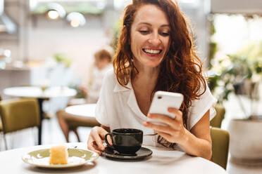 Junge Frau, die auf ihrem Smartphone in ihren Lieblings-Social-Media-Apps surft, während er in einem Café einen Kaffee genießt. Kaukasische Frau mit rotem Haar, die ihre eigene Gesellschaft in einem Café genießt. - JLPSF30099