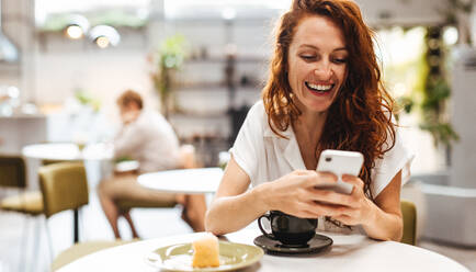 Kaukasische Frau tippt auf ihrem Telefon und schickt Textnachrichten an ihre Freunde, während sie ein heißes Getränk in der gemütlichen Atmosphäre eines Cafés genießt. Glückliche Frau, die ein Kaffee-Date hat. - JLPSF30100