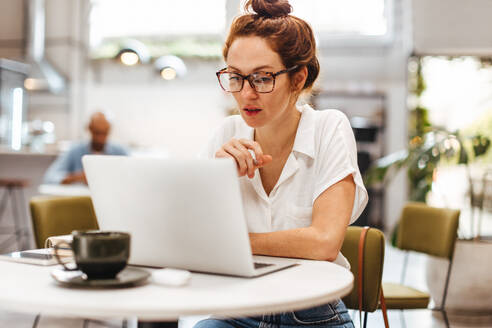Kaukasische Unternehmerin arbeitet an ihrem Laptop in einem gemütlichen Restaurant. Frau mit roten Haaren sitzt in einem Café und liest durch ihre Brille über ihr nächstes Geschäftsprojekt. - JLPSF30106