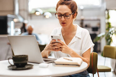 Focused female business professional checks her mobile phone for messages as she works remotely from a cozy coffee shop, managing her work tasks and communications with ease. - JLPSF30107