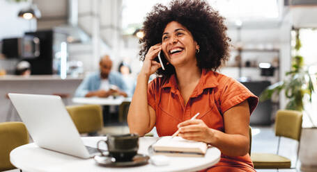 Professional businesswoman managing her appointments and communicating with clients over the phone. Happy young woman working remotely from her favourite cafe and work spot. - JLPSF30110