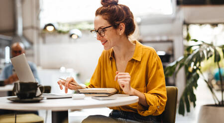 Eine engagierte Studentin arbeitet in einem gemütlichen Café an ihrer Dissertation. Eine glückliche kaukasische Frau sitzt an einem Tisch in einem Café und recherchiert mit einem Laptop für ihr Studium. - JLPSF30113