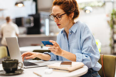 Junge Frau benutzt ihre Kreditkarte und ihren Laptop für Online-Banking, während sie in einem Café sitzt. Geschäftsfrau, die elektronische Zahlungen für ihre freiberufliche Tätigkeit tätigt. - JLPSF30114