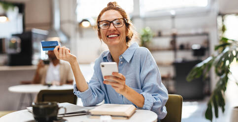 Portrait of a happy woman using her mobile phone and credit card for online shopping in a café. Red haired young woman making electronic payments on her mobile banking app. - JLPSF30115