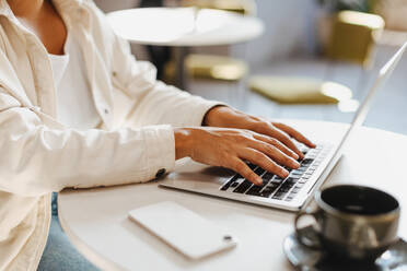 Business woman using her laptop to type an email to a client in a cafe. Young female professional sitting at a table in a coffee shop, engaged in remote work. - JLPSF30117