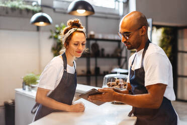 Zwei Servicefachleute im Gastgewerbe verwenden ein Tablet, um Bestellungen effizient zu verwalten und die Kundenzufriedenheit zu gewährleisten. Technisch versierte Kellner arbeiten als Team in einem Café. - JLPSF30126