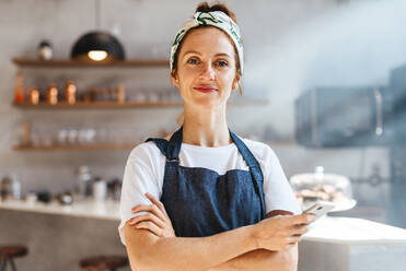 Caucasian coffee shop owner standing in her establishment, embracing technology with her smartphone and using it to streamline operations and provide a modern, efficient experience for her customers. - JLPSF30139