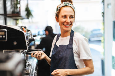 Female coffee shop employee using a coffee machine to prepare coffee for customers. Happy barista using her coffee brewing skills to provide excellent service and hospitality in a cafe. - JLPSF30142